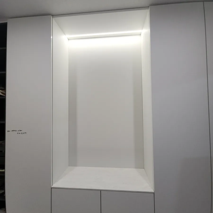 흰 수납장 사이 오목한 벽감 윗부분에 LED 조명이 켜져 있고 하단에 서랍장이 있는 모습
